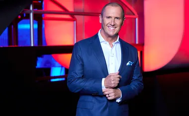 Larry Emdur, wearing a white shirt and blue suit, stands on the set of The Chase