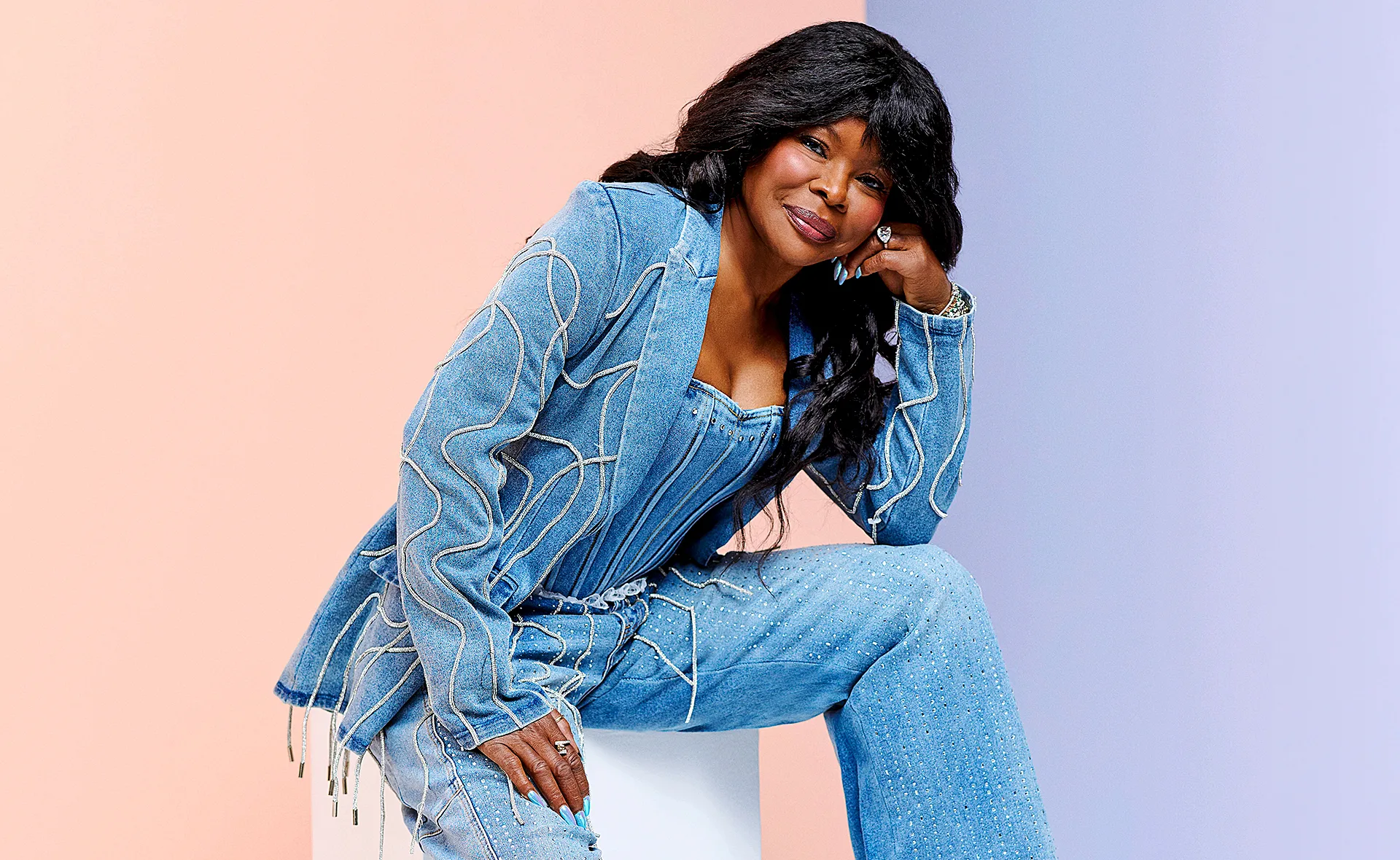 Marcia Hines, in a blue pantsuit, sits on a white block with her head resting on her hand
