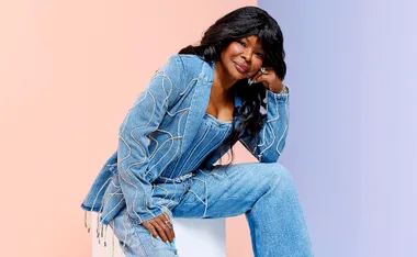 Marcia Hines, in a blue pantsuit, sits on a white block with her head resting on her hand