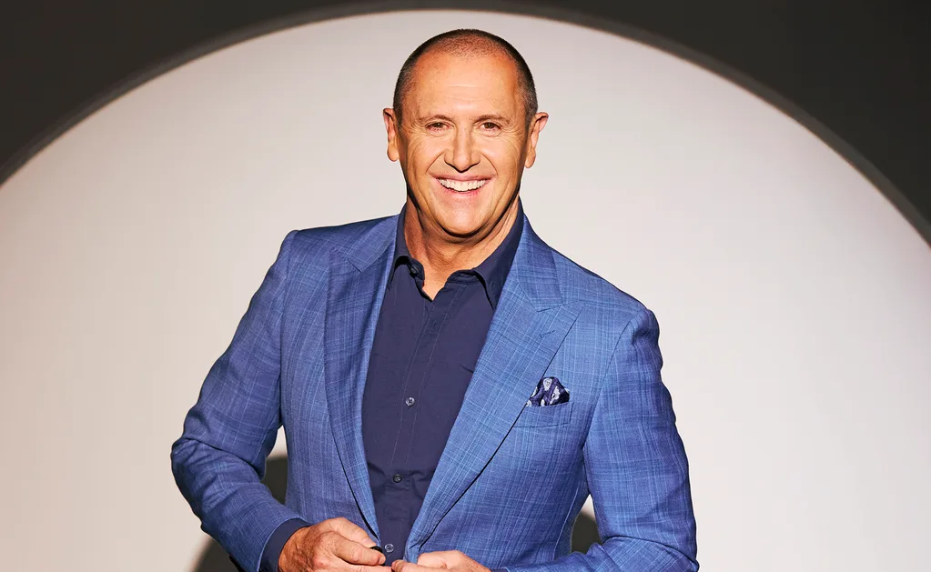 Larry Emdur, in a blue shirt and blue suit, smiling, stands in front of a white and black background.