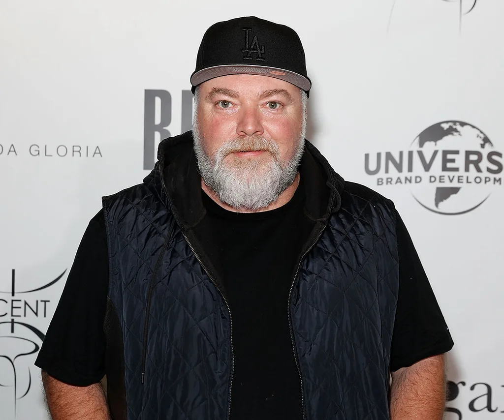 Kyle Sandilands poses in front of a media wall in a black t-shirt, puffer vest and cap.