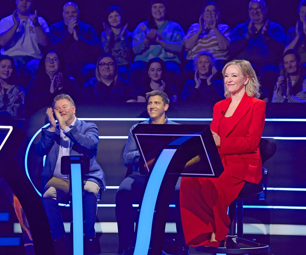 Rebecca, in a red suit, sits on the set of Millionaire Hot Seat, with two other people in the background