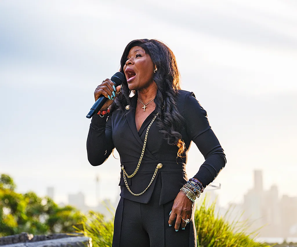Marcia Hines, wearing a dark outfit with chains, sings inot a microphone, outside, with the sun behind her