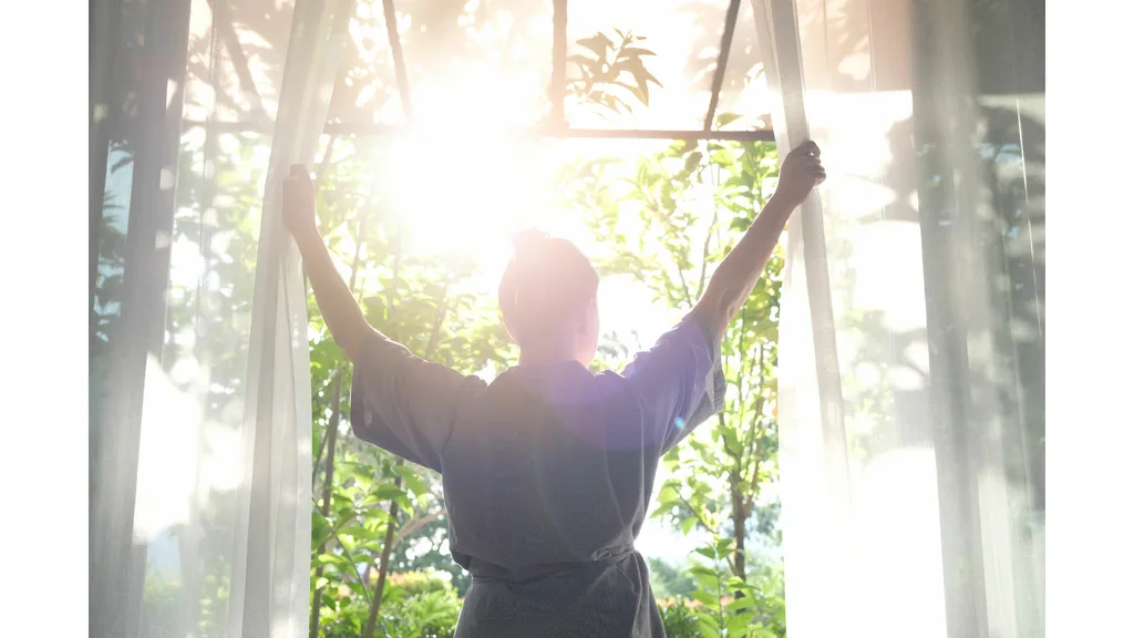 A woman stands at a window in a robe and opens the curtains as sunlight filters in.