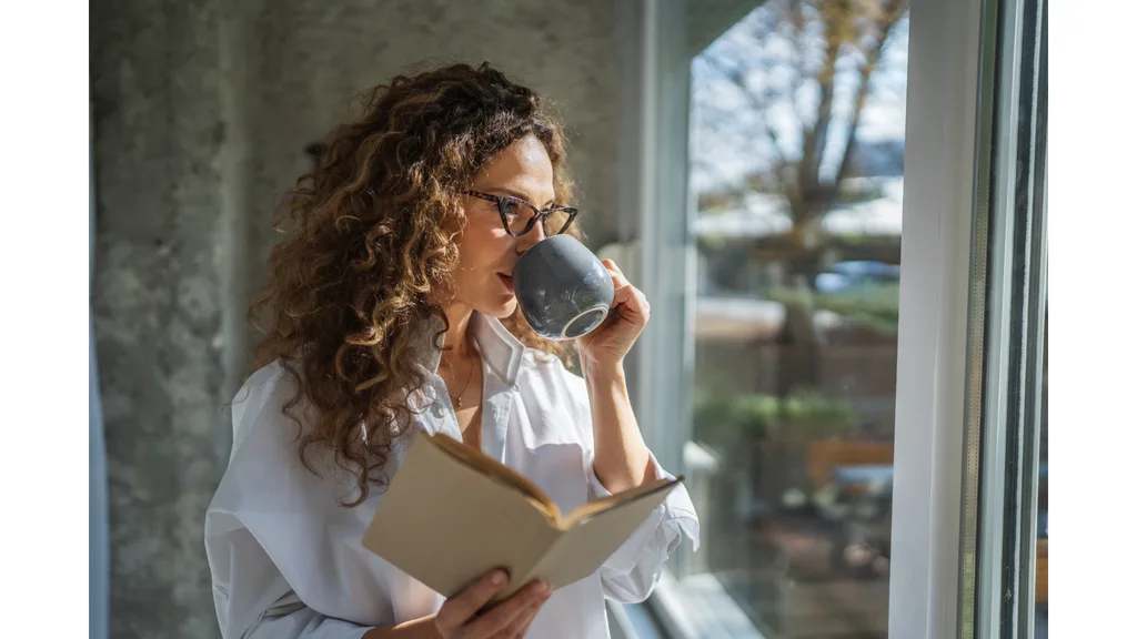 A woman sips from a mug while holding an open book and gazing out the window.