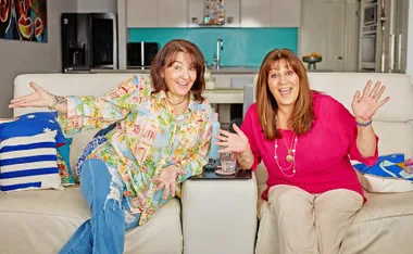Anastasia, in a patterned top, sits on a lounge chair, leaning towards Faye, in a red top, who is also sitting on a lounge chair