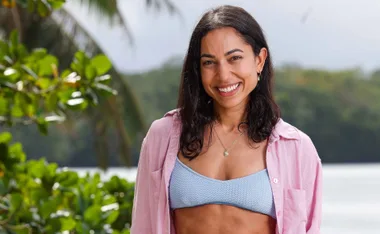 Brooke, wearing a blue bikini top and a pink shirt, stands in front of a tree and the sea, smiling