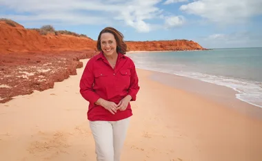 Lisa Millar, in a red top, walks along a beach with red cliffs behind her