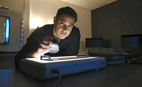 Marc Fennell holds a magnifier and looks up, in a room full of shadows