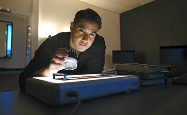 Marc Fennell holds a magnifier and looks up, in a room full of shadows