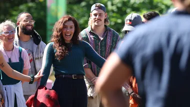 Eliza, wearing a green jumper, stands among a group of people on Survivor