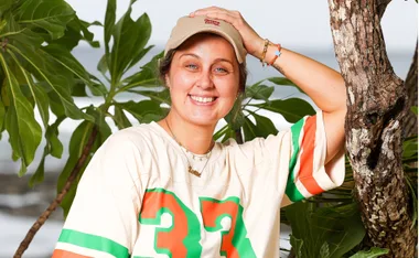 Lottie, in a t-shirt with the number 37 on it, holds her cap onto her head, with vegetation and the sea in the background