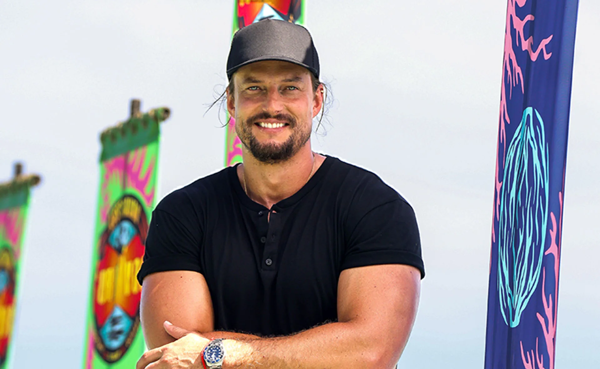 David Genat, wearing a cap and a black t-shirt, stands with his arms folded, smiling, in front of Survivor flags