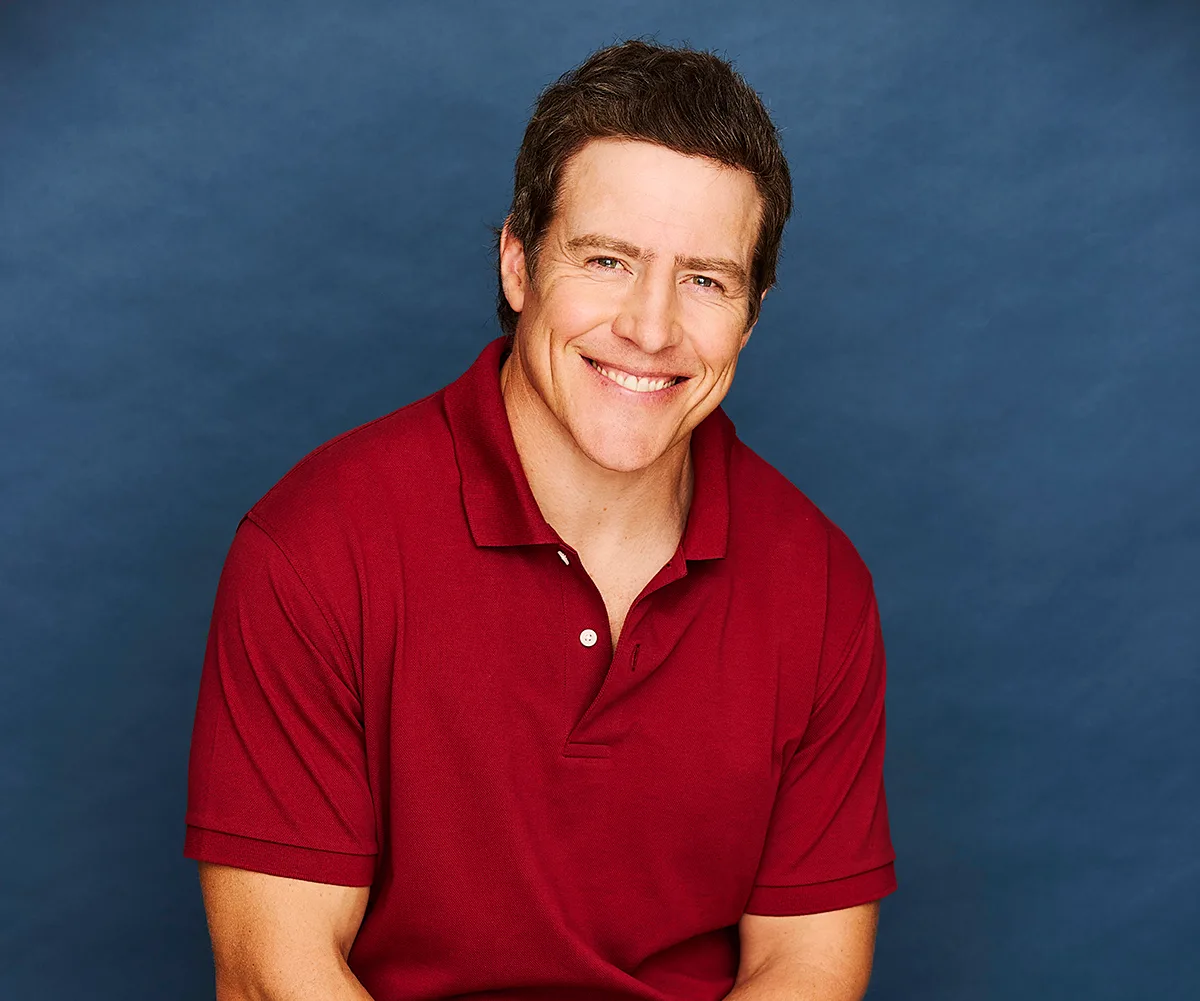 Stephen Peacocke, in a red shirt, sitting in front of a blue background, smiles 