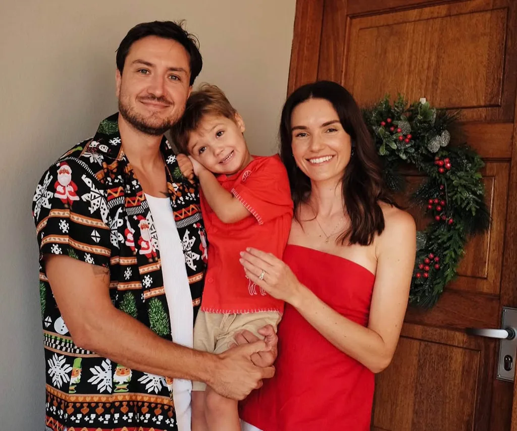 Amanda, in a red dress, stands with her husband, in a patterned shirt, who is holding her young son, all three in front of a Christmas wreath