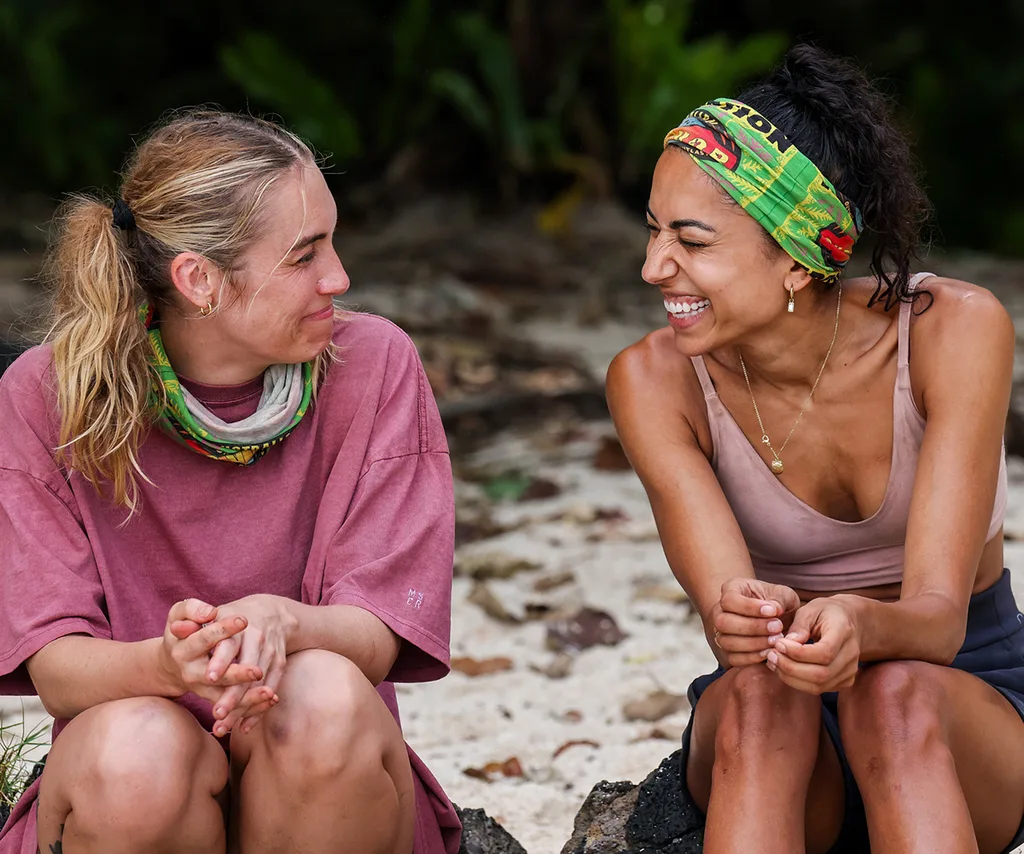 Brooke, wearing a Survivor buff on her head, sits next to Faith, smiling at her, on the beach in Samoa