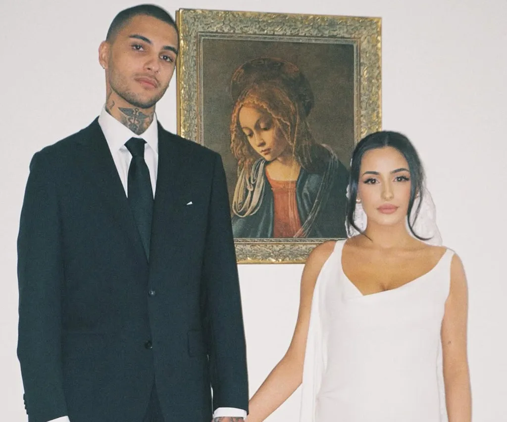 Tre, in a suit and tie, holds hands with his wife Victoria, in a white dress, both standing in front of a painting