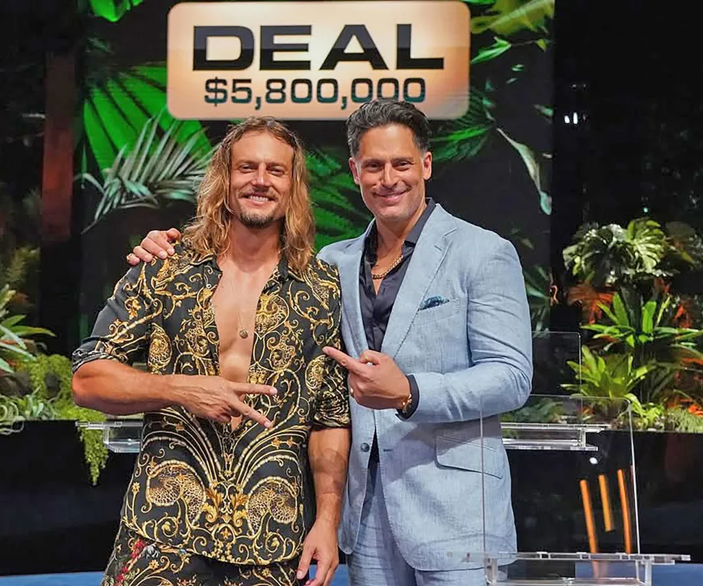 David, with a patterned shirt unbuttoned, stands next to Joe Manganiello, wearing a jacket, with a sign behind them saying "DEAL $5,800,000"