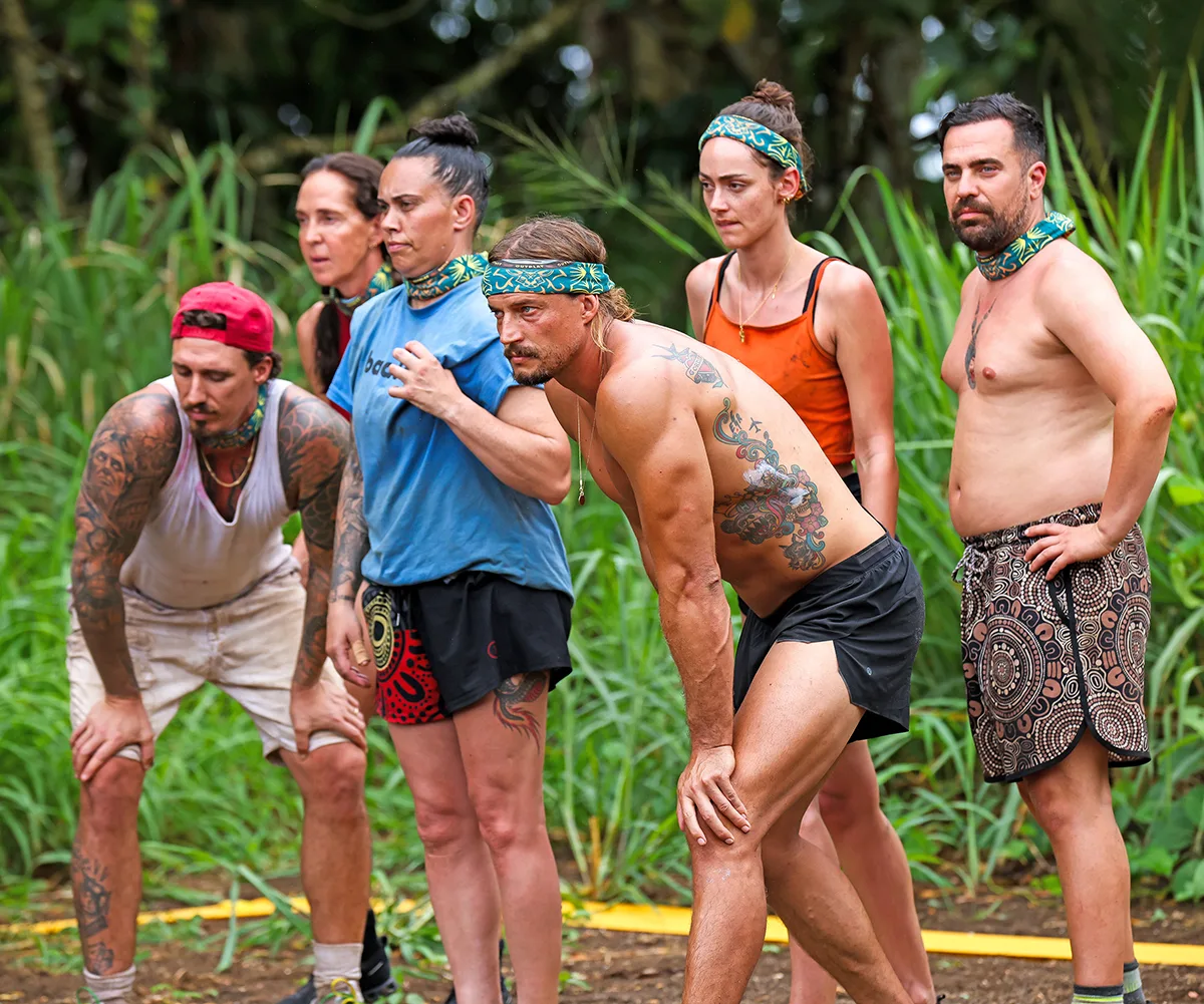 David, shirtless, wearing a buff as a headband, leans forward intently in Samoa, other Survivor players standing behind him
