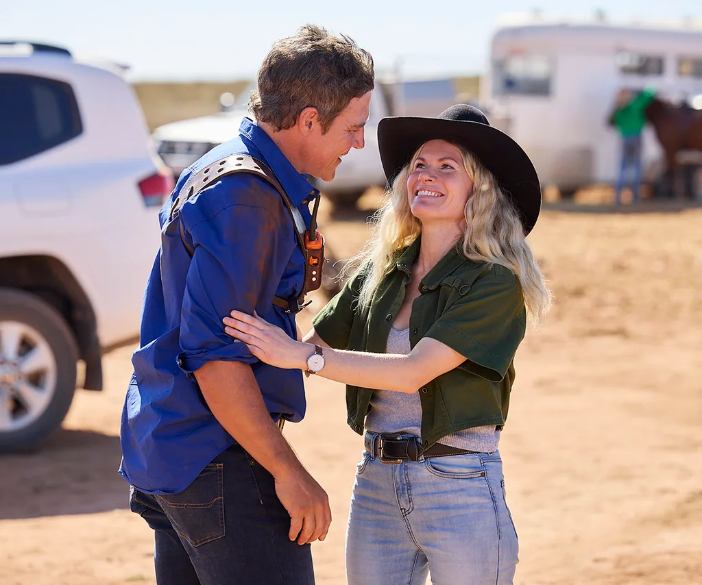 Bonnie, wearing a hat, affectionately touches Stephen, in a blue shirt, as the two of them play Brax and Ricky in an outback setting