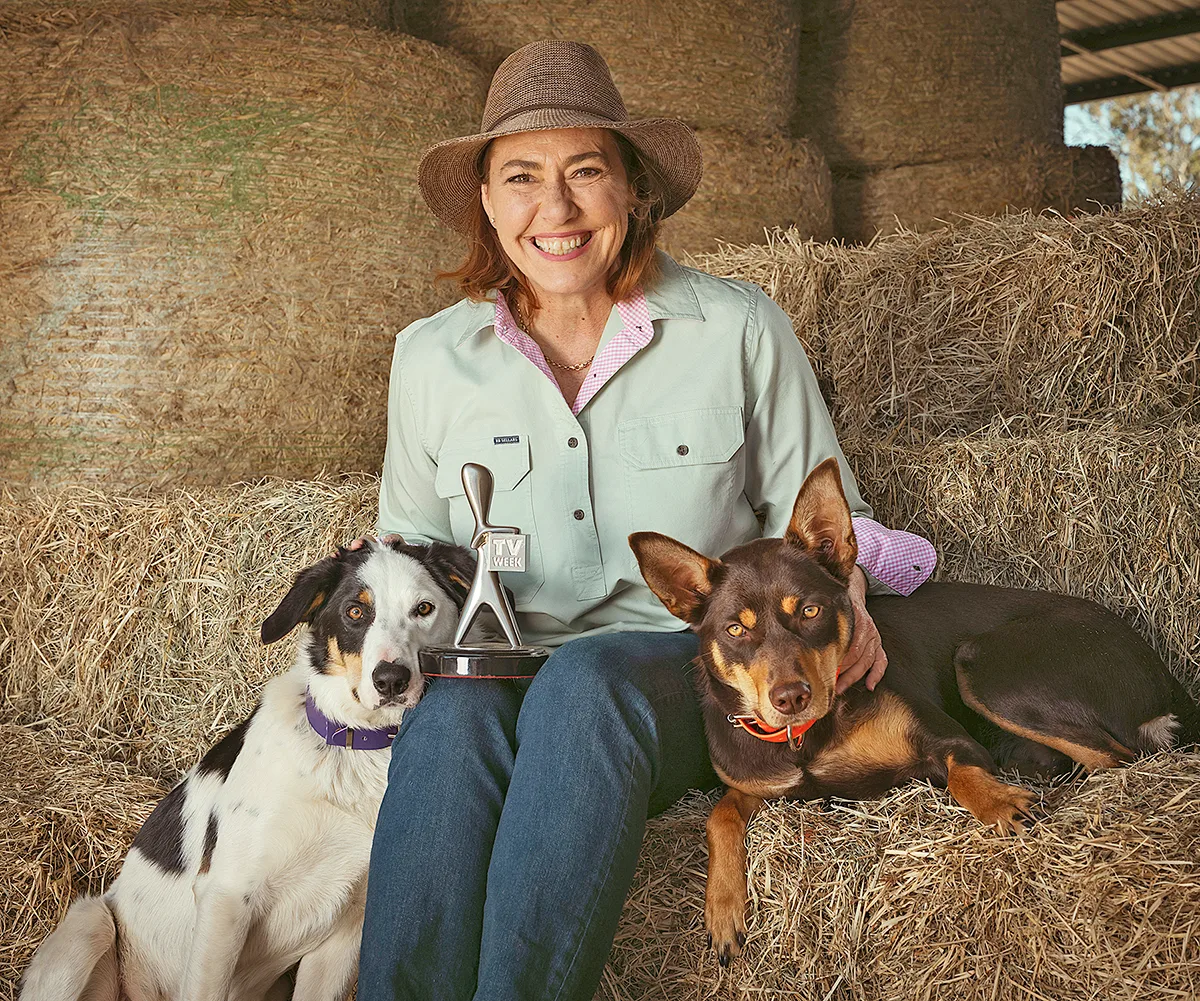 Lisa, wearing a hat, sits on a bale of hay, with a young collie on one side and a young kelpie on the other
