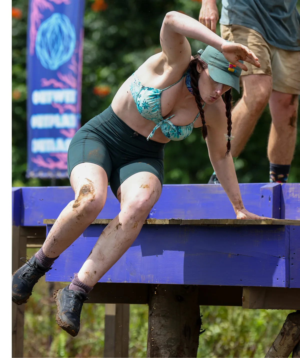 Eliza, wearing a bikini top and shorts, jumps off a wooden platform in a challenge