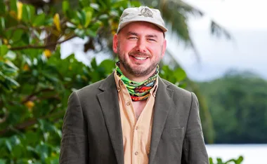 Mark, wearing a jacket and a cap, and standing in front of a tree on a beach in Samoa