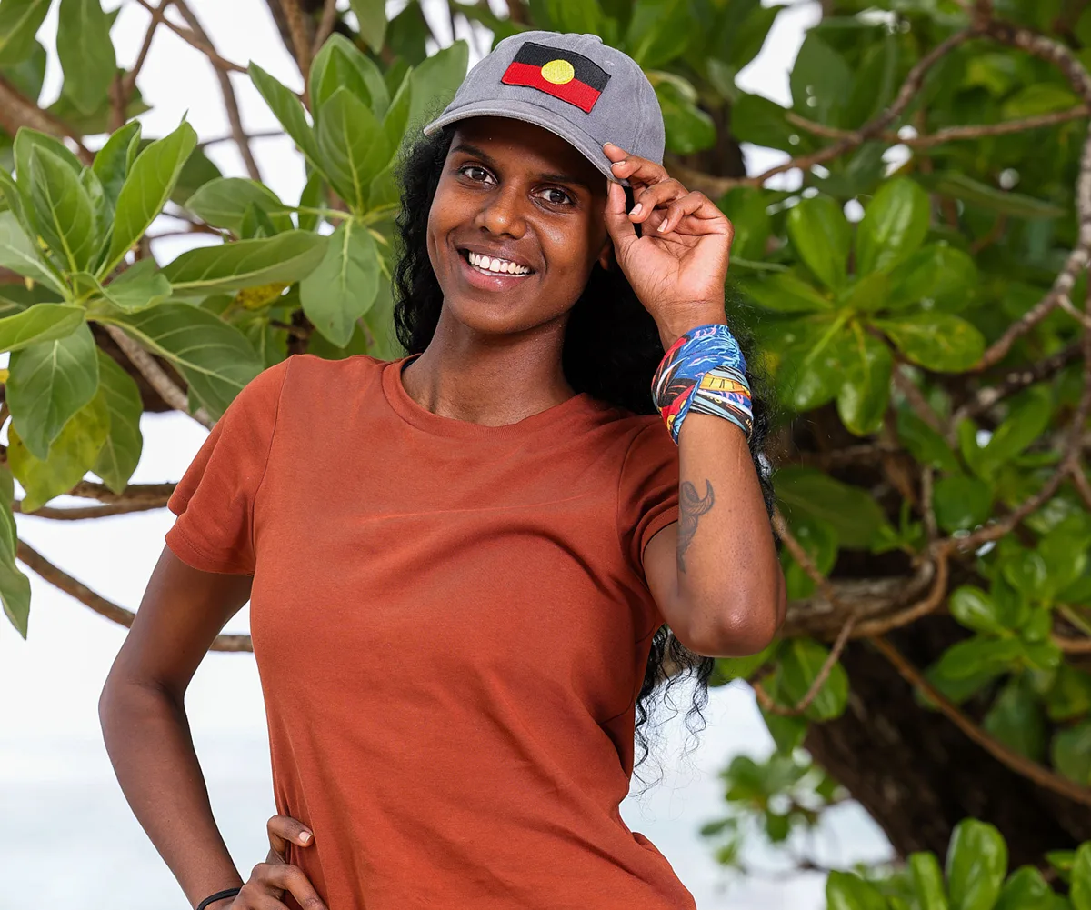 Aisha, wearing a brown top, and a cap with the Indigenous flag on it, stands in front of a tree
