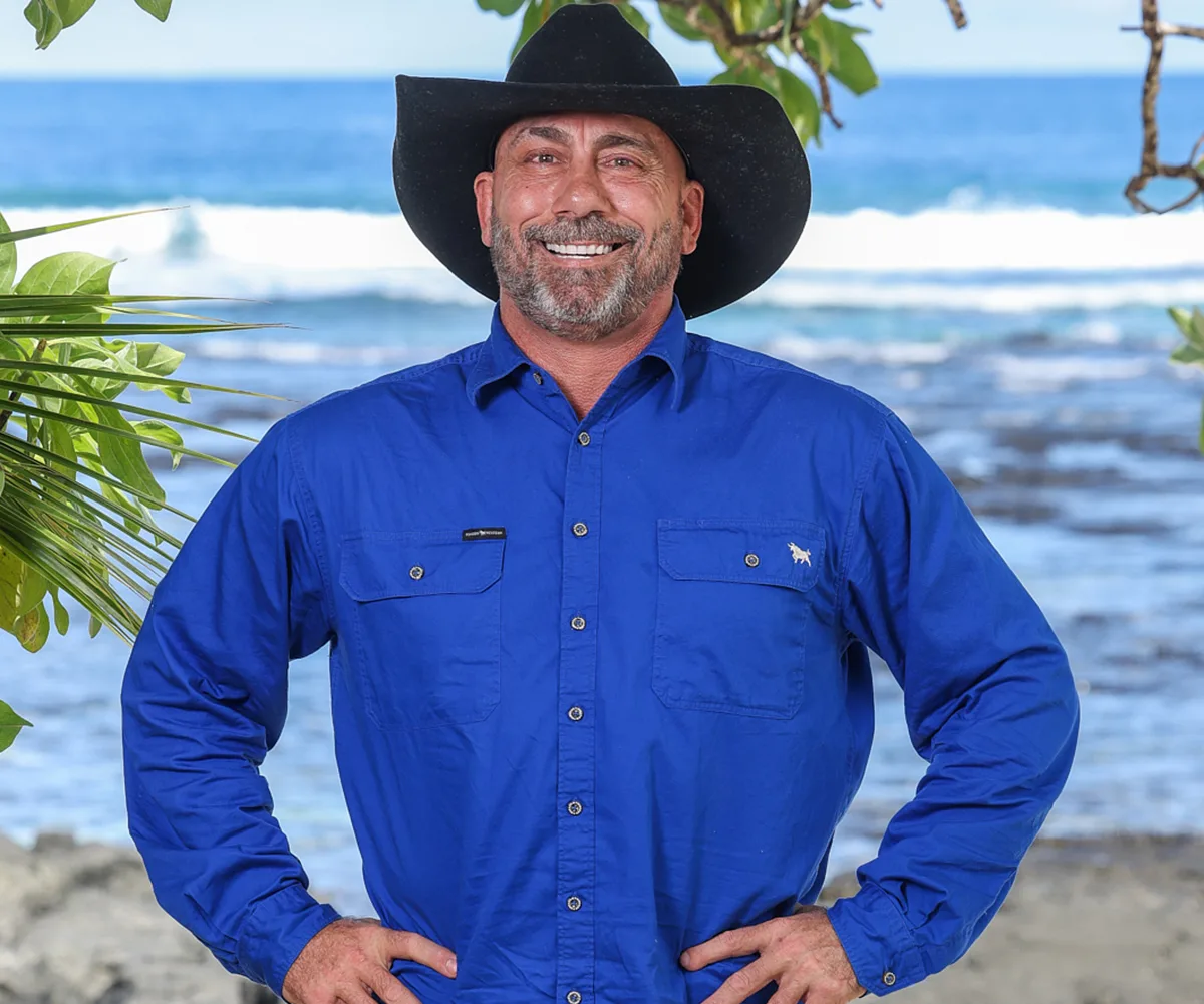 Don, with a blue shirt and a cowboy hat, stands with his hands on his heaps, the sea in the background