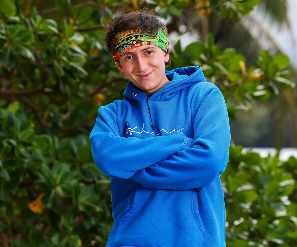 Tez, wearing a blue hoodie and a bandana around his head, stands with his arms folded, on a beach in Samoa
