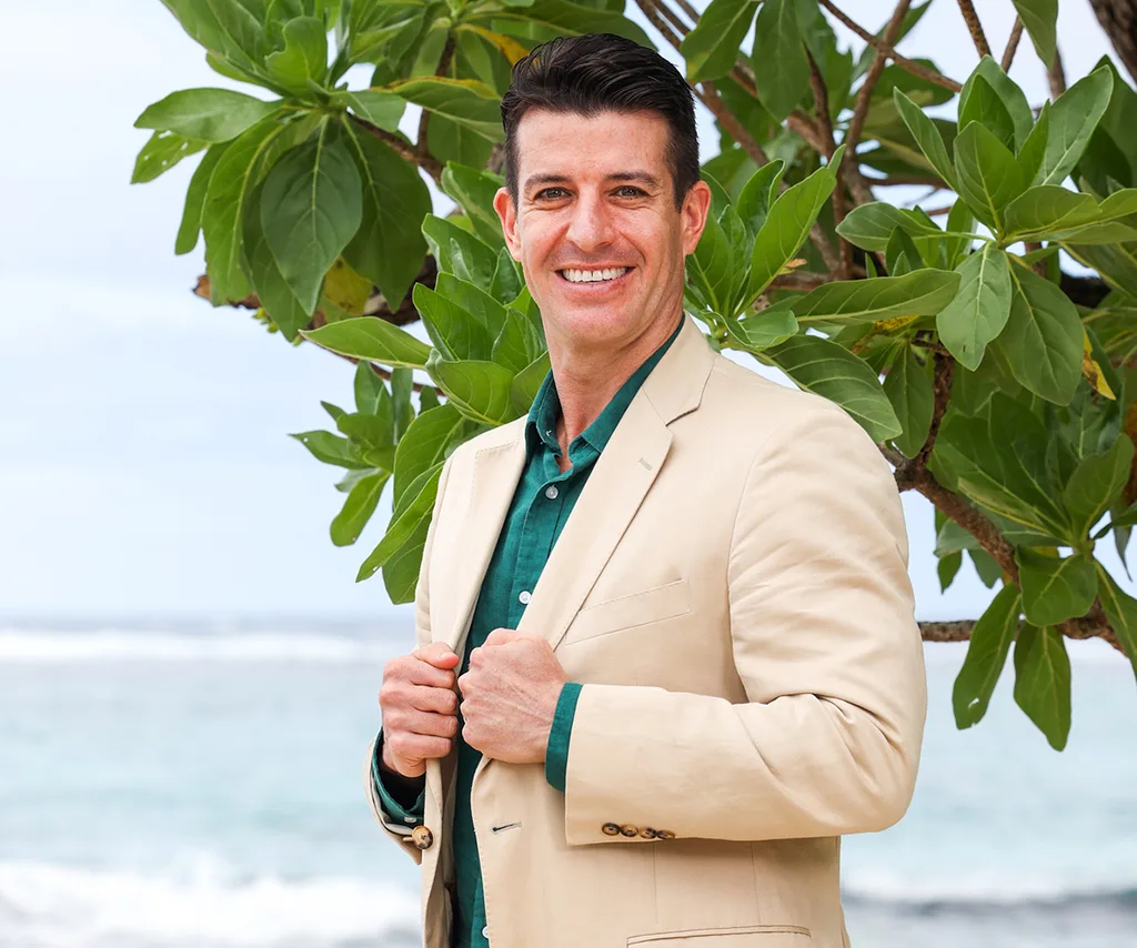 Richard, in a green shirt and cream jacket, stands with his hands holding his lapels, in front of a tree on a beach in Samoa