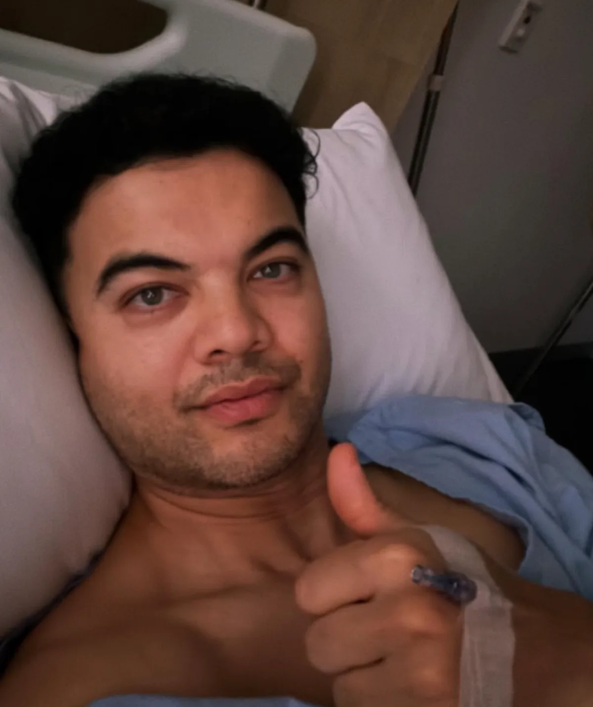 Guy lies in a hospital bed, giving a thumbs-up