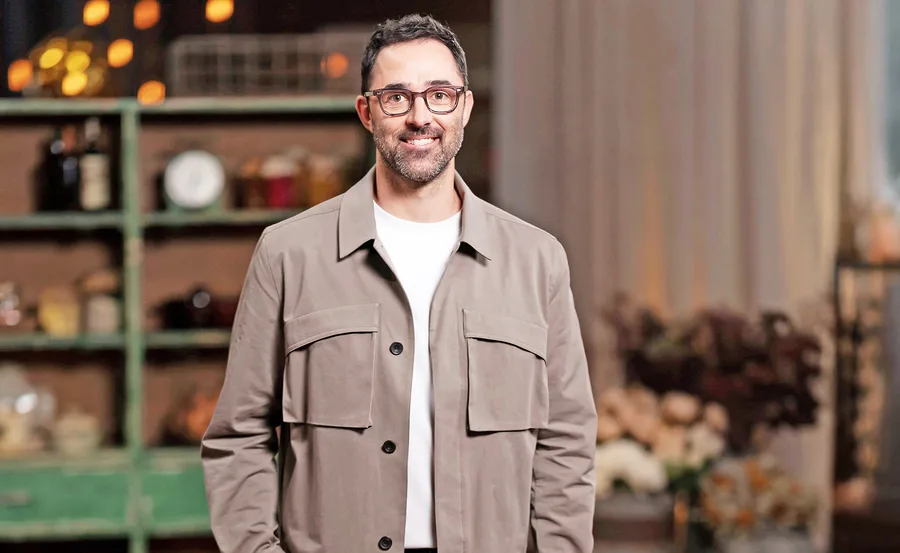 Andy Allen dressed in a beige jacket with white t-shirt underneath, smiling in the MasterChef Australia kitchen.