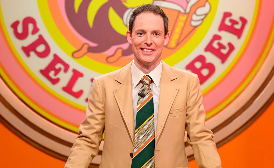 Guy, wearing a striped tie and a salmon-coloured jacket, stands on the set of his Spelling Bee