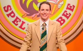 Guy, wearing a striped tie and a salmon-coloured jacket, stands on the set of his Spelling Bee