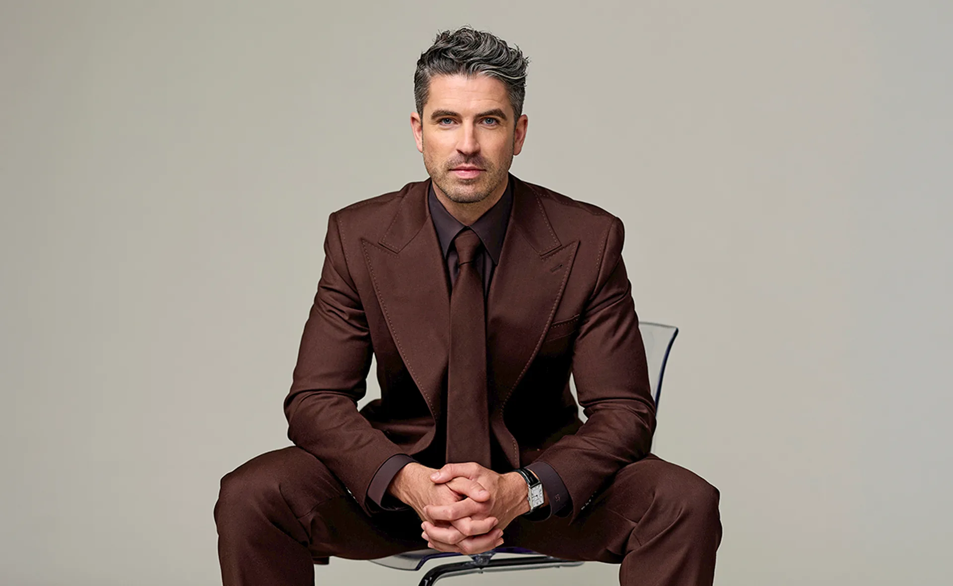 Scott Tweedie, host of Australian Idol, sitting on a chair in a brown suit.