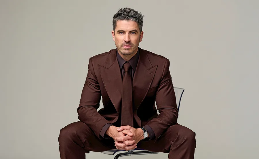 Scott Tweedie, host of Australian Idol, sitting on a chair in a brown suit.