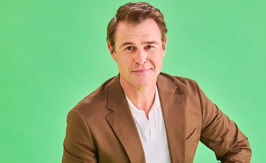 Rodger, wearing a brown suit, sits in front of a green background