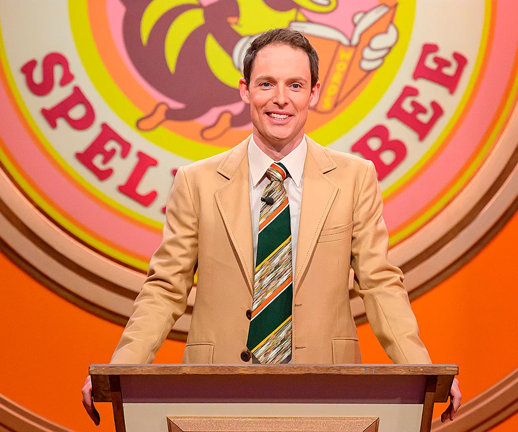 Guy stands behind a podium on the set of Spelling Bee