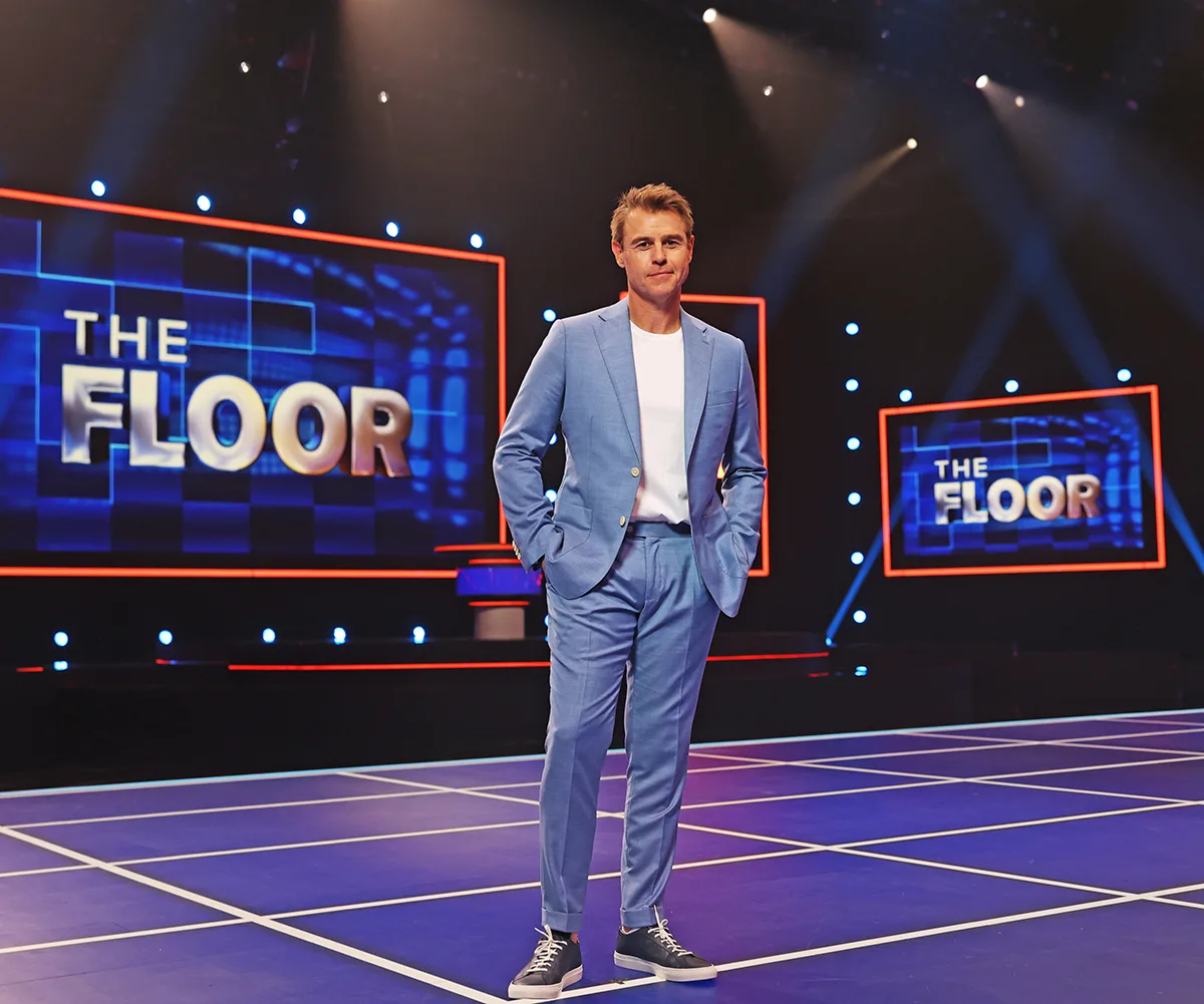 Rodger, wearing a blue suit, stands on the set of The Floor.