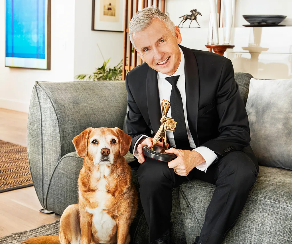 Bruce, in suit, sits on a couch, holding a Logie, with his brown and white dog Frankie sitting next to him