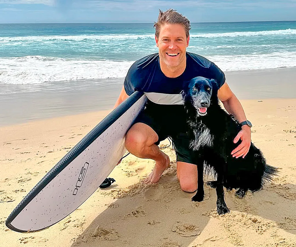 Dr Chris Brown on the beach holding a surf board and posing with his dog Buzz.