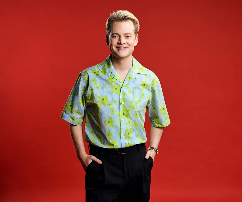 Joel Creasey, wearing a patterned shirt, stands in front of a red background