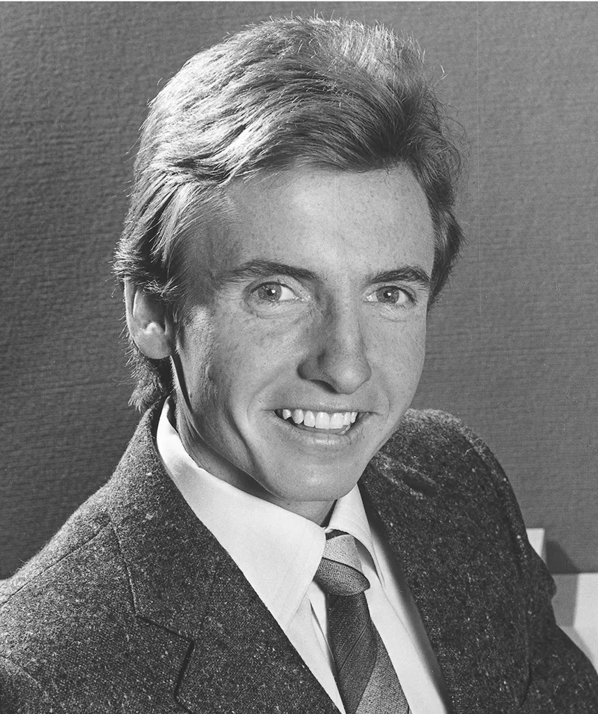 A black-and-white photo of a young Bruce McAvaney, in a suit and tie, smiling at the camera
