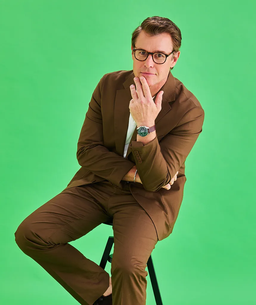 Rodger, wearing glasses and a brown suit, sits with his hand on his chin in front of a green background