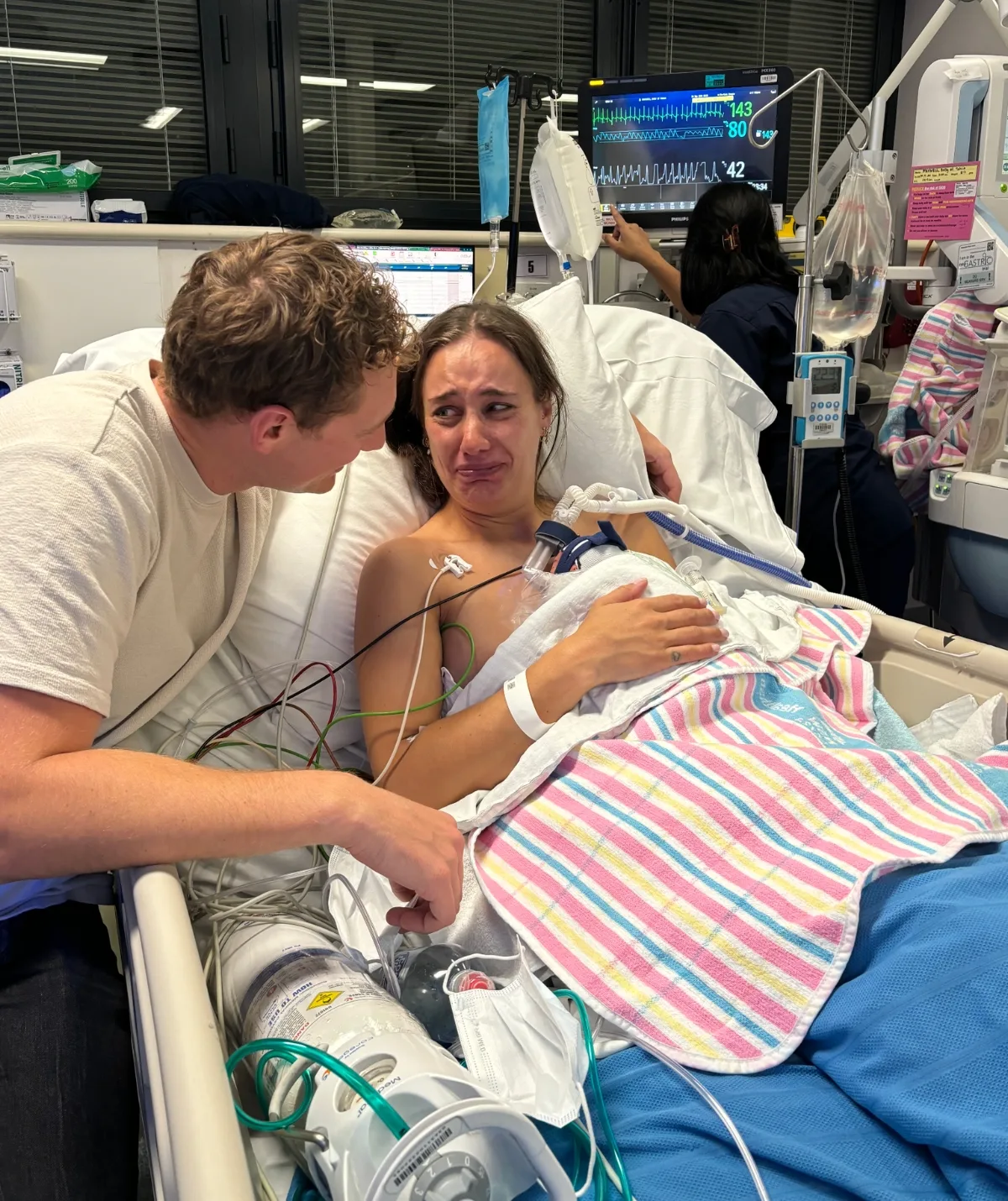 A man looks over a woman lying in a hospital bed who is crying and has a baby on her stomach.