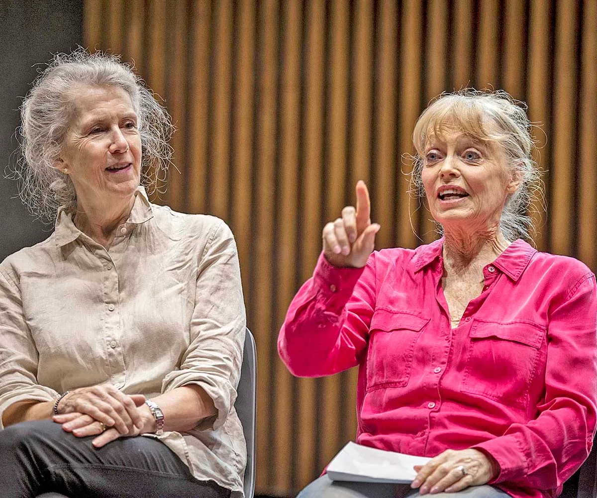 Belinda raises her finger in the air at a rehearsal, sitting next to Debra