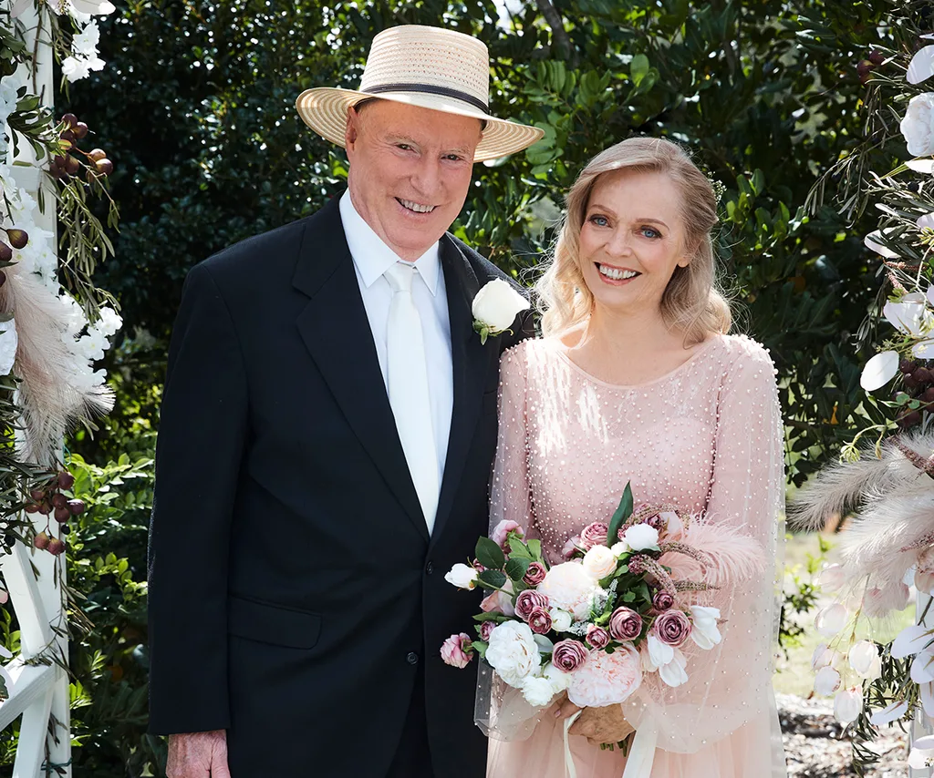 Belinda, as Martha, in a wedding dress, stands next to Ray Meagher, as Alf, in a wedding suit, on the set of Home And Away