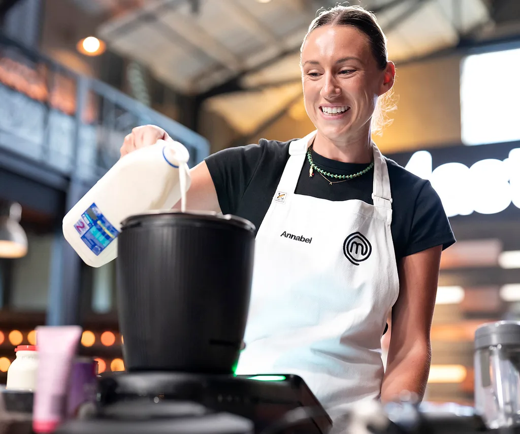 Annabel Lloyd in the MasterChef Australia kitchen smiling pouring milk into a pot.