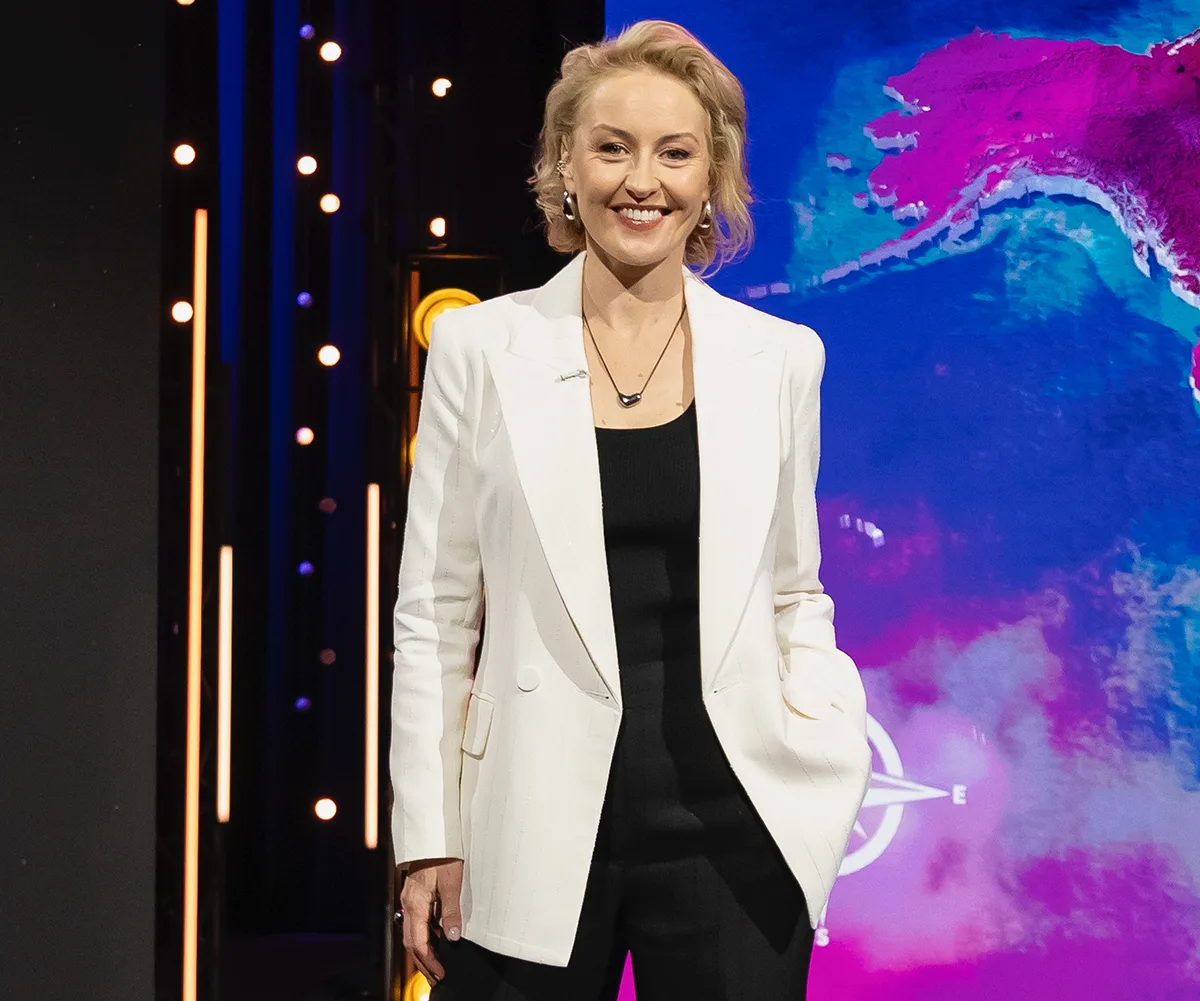Zan Rowe, wearing a white jacket, stands on the set of Race Around The World